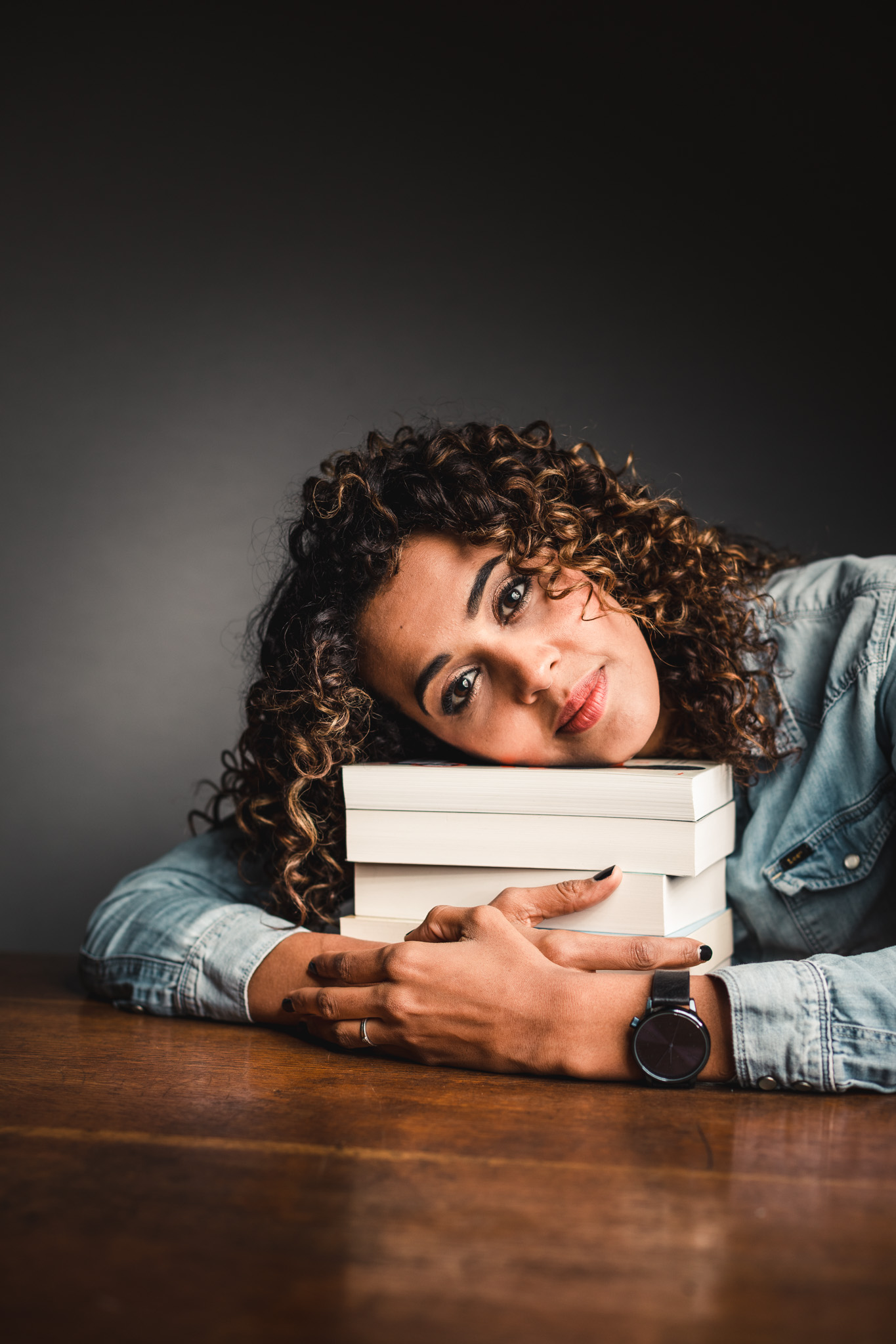 Tatyana Beloy zit aan tafel, waar een stapel boeken ligt. Haar hoofd rust op de stapel, die ze met beide armen omarmt. Ze kijkt recht in de lens en glimlacht. De achtergrond is grijs tot zwart.