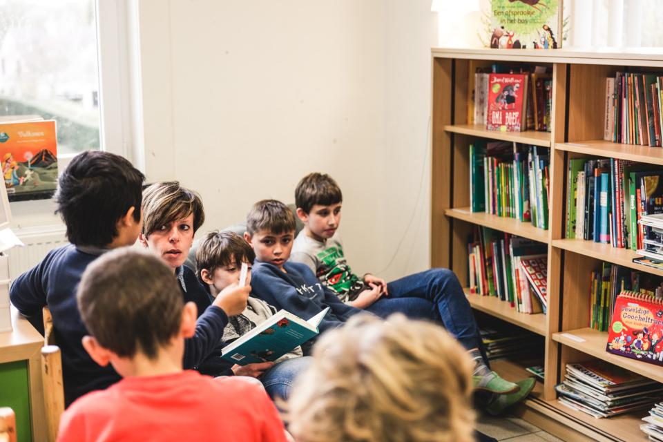 Leerlingen zitten op een rij. In het midden leest een juf voor. Rechts staat een boekenkast