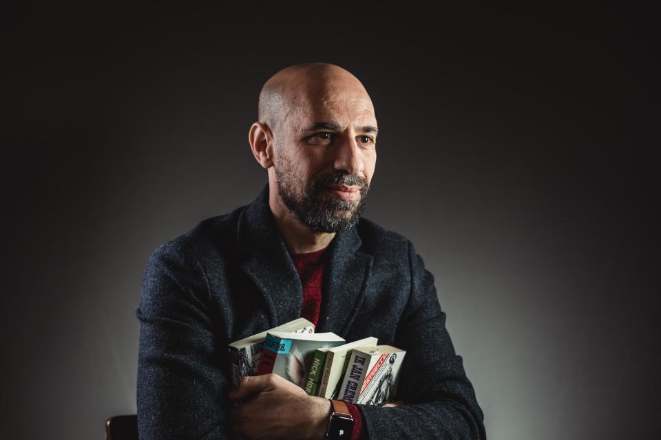 Portret van auteur Abdelkader Benali. Hij omklemt vier boeken in zijn armen en kijkt weg van de lens. De achtergrond is grijs.