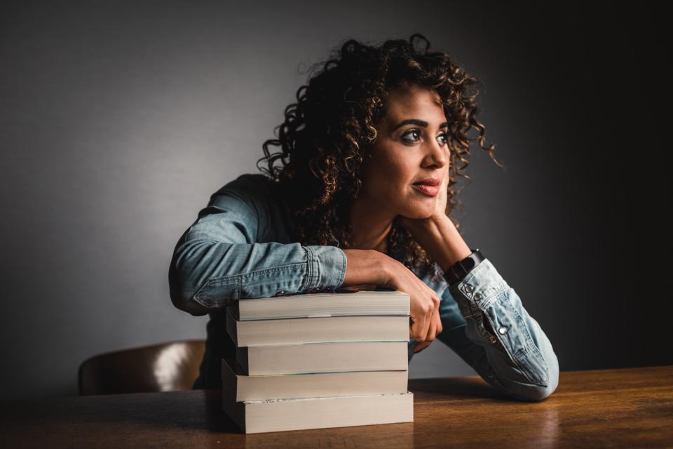 Tatyana Beloy zit aan tafel met een stapel boeken voor zich, waarop haar rechterarm op leunt. Haar hoofd leunt op haar linkerhand. De achtergrond is grijs.