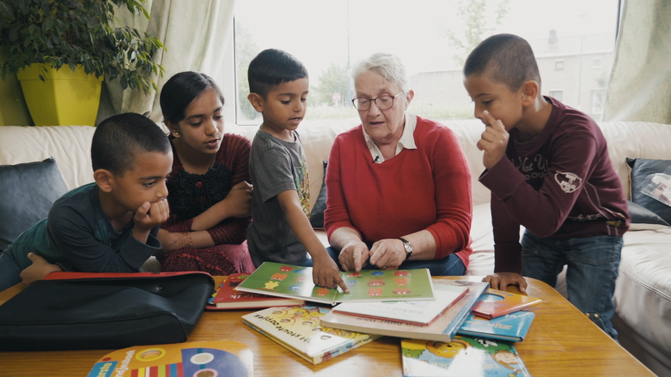 Vier kinderen zitten aan de salontafel, een vrijwilligster leest voor. Op de salontafel liggen verschillende prentenboeken.