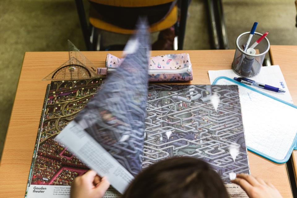 Een leerling slaat de bladzijde van een groot prentenboek om dat op een lessenaar ligt. De foto is genomen van bovenaf. Op de lessenaar liggen geodriehoeken, een etui, schrijfgerei en een schrift.